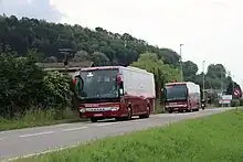 Photographie en couleurs de deux autocars rouges sur une route bordée par un pré vert. En arrière-plan se détachent une haie et le toit de quelques habitations.