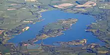 Photo aérienne d'une étendue d'eau entourée de champs verts et de petits villages. Un îlot de forme ovale se dresse au milieu de l'eau, relié à la terre ferme par un isthme en bas de l'image