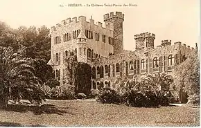 Château néo-médiéval avec tours crénelées, échauguette et palmiers ; nombreuses fenêtres en façade avec volets ouverts cintrés.