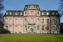 une imposante bâtisse rose de style rococo à la façade percée de douze travées sur deux niveaux et toit mansardé surplombe une pelouse entourée de hauts arbres