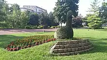 Au milieu d'un espace de pelouse verte, près d'un chemin piéton en asphalte rose, se dresse une sculpture végétale représentant un paon. Placée sur une petite butte maintenue par un mur de pierre de quelques marches, le corps de l'animal est constitué d'un seul arbuste taillé en forme de corps, cou et tête. Un cône métallique vient symboliser le bec. La queue de l'animal, posée au sol derrière lui, est composée d'un parterre de fleurs orange et rouges sur le pourtour.