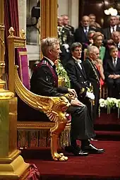 Photographie de profil  d'un homme aux cheveux blancs assis sur un fauteuil aux accoudoirs dorés