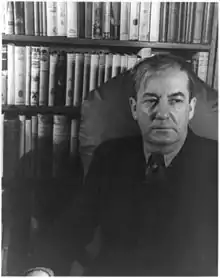 Homme assis devant rayonnages de livres, 50&nbsp;ans environ, cheveux avec raie, costume-cravate.