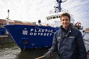 Simon Bernard pose devant le bateau Plastic Odyssey, mis à l'eau derrière lui.