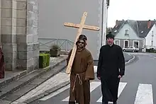 Deux hommes sont déguisés. L'un est habillé d'une robe traditionnelle de moine et de lunettes de soleil, portant sur son dos une grande croix en bois. Le deuxième porte une soutane noire, des moustaches de chat sont maquillées sur son visage.