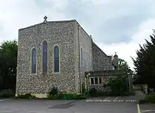 Photo d'un bâtiment à la façade de pierre grise imposante, percée de trois fenêtres en ogive, surplombé d'une croix chrétienne