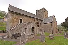 Photo d'un bâtiment allongé en pierre ocre, flanqué d'une grande tour et d'un porche à droite. Des stèles funéraires sont visibles au premier plan