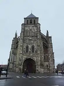 Tour-porche présentant une couleur grisâtre. En haut de la tour, toiture grise ordinaire avec une croix surmontée d'un coq. Ciel gris.
