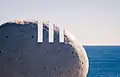 Photo agrandie du coin supérieur droit de l'une des pierres commémoratives. Trois entailles droites y sont incrustées, représentant le chiffre 111. À l'arrière-plan, la mer baigne au pied d'un ciel blanc de soirée.