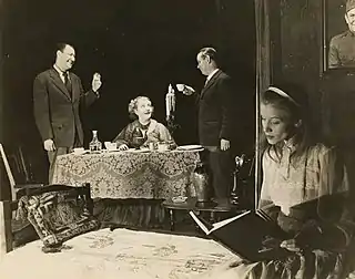 À l'arrière-plan 2 hommes portent un toast à une femme assise derrière une table, tandis qu'une autre femme lit un livre à l'arrière-plan à droite.