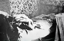 Photographie en noir et blanc d'un lit défait et désordonné. Une pistolet est posé sur le lit.