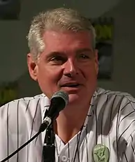 Photo en buste d'un homme avec les cheveux gris, portant une chemise blanche rayée et parlant dans micro devant lui.