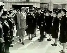 Photographie en noir et blanc représentant deux femmes et trois hommes entourés de  militaires vêtus de costume sombre.