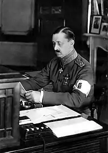 Photo en noir et blanc d'un homme moustachu lisant assis à une table, il porte un uniforme militaire et un brassard blanc au bras gauche