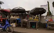 dans une rue, deux étals en bois, abrités par un parasol, adossés à un mur, présentant du pain pour l'un et des fruits pour l'autre