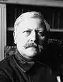 Photo en noir et blanc d'un homme moustachu au visage rond, aux cheveux blancs.