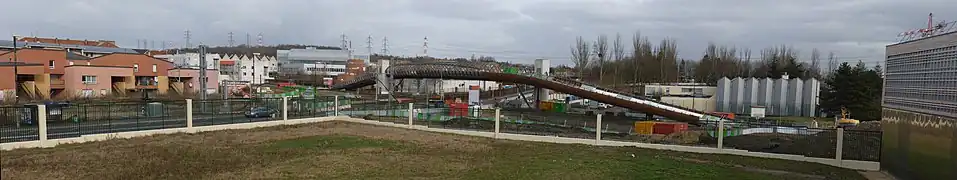 Le premier ouvrage d'art construit sur le Tram Express Nord : la passerelle de Villetaneuse-Université, photographiée pendant son chantier de construction, desservant la gare construite dans un second temps à cheval sur les voies, et qui relie l'Université Paris-XIII, dont on voit le gymnase universitaire à droite du cliché, et le quartier du Centre-ville, à gauche. Au fond à droite, le Centre nautique Jacques-Duclos, démolit depuis.