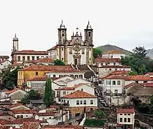 Photographie en couleur. Vue panoramique de la vieille ville d'Ouro Preto avec des maisons blanches au toit de tuiles rouges surplombées d'une église blanche en haut d'une colline.