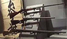 Photographie d'un présentoir de musée sur lequel sont placés trois fusils de forme et de taille différentes, la crosse à gauche et le canon à droite