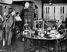 Photographie en noir et blanc d'un plateau de cinéma, le réalisateur se tenant debout à gauche, avec un technicien, entre des projecteurs, l'acteur à la tête bandée se tenant à droite, assis à une table sur laquelle sont posés divers objets.