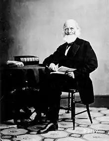 La photographie noir et blanc montre un homme âgé dans un costume noir. Il porte une barbe blanche imposante, et tient un livre ouvert, assis sur une chaise. Il est accoudé à une table sur laquelle repose un épais livre noir et une liasse de papier.