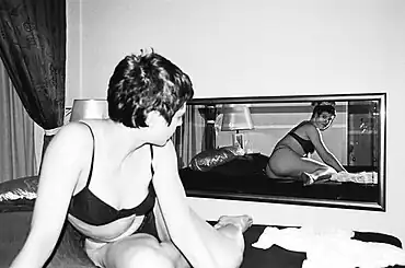 Une jeune femme blanche, en sous-vêtements noirs, aux cheveux bruns et courts, se tient à moitié allongée sur un lit. Elle regarde un miroir derrière elle, qui reflète son image.