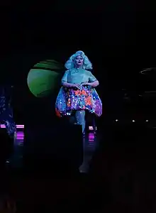Kim Chi est sur scène. Son haut et sa perruque sont bleu-vert irisé ; sa jupe a un motif floral multicolore.