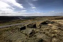 Des collines recouvertes de pierres et d'herbes apparaissent sur la photo. En arrière-plan, un ciel nuageux est présent. Une route sépare les collines et une rivière coule au fond.